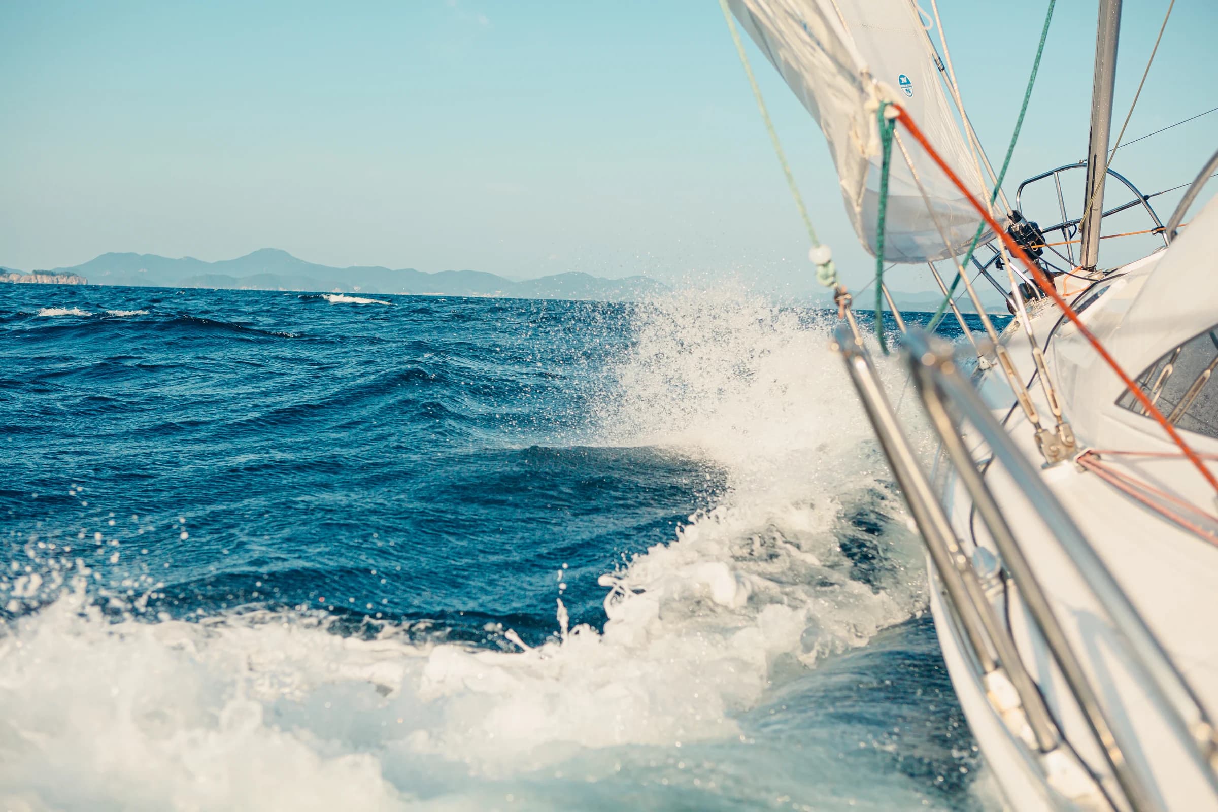 A wave breaks alongside a sailing yacht on a deep blue sea.